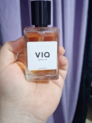 VIQ Idyllic Eau de Parfum - En stilren parfymflaska från VIQ med doften Idyllic. Flaskan är rektangulär i genomskinligt glas med en svart fyrkantig kork och en vit etikett med svart text. Innehållet har en varm bärnstensfärgad ton. Perfekt för dig som gillar minimalistisk design.