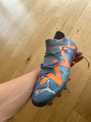 Blå-orange fotbollssko från Puma - Snygg fotbollssko från Puma i blått med orange detaljer och vita loggor. Skon har en strumpliknande krage, snörning och greppvänlig sula med dobbar för bra fäste på planen. Tillverkad i syntetmaterial för lätt känsla och bra bollkontroll. Nypris 1699 använda 7 gånger