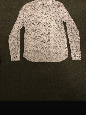 Vit blommig skjorta med knappar - Snygg vit skjorta med små blommiga mönster i blått och svart. Skjortan har lång ärm, klassisk krage och knäppning framtill. Perfekt för dig som vill ha en stilren men ändå lekfull look.