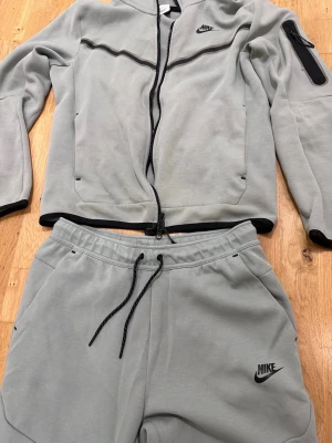 Grått Nike loungewear-set M - Säljer ett grått loungewear-set från Nike i mjukt material. Setet består av en zip-hoodie med svarta detaljer och fickor samt matchande byxor med dragsko och sidofickor. Perfekt för chill dagar hemma eller när du vill vara bekväm.
