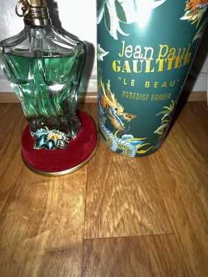 Jean Paul Gaultier Le Beau Paradise Garden - Har använt ungefär 10 ml av den Unik parfymflaska formad som en manlig torso med grön vätska och guldfärgad spraydel. Kommer i en dekorativ cylinderförpackning med tropiskt mönster och texten 'Le Beau Paradise Garden'. Perfekt för dig som gillar snygg design och fräscha dofter.