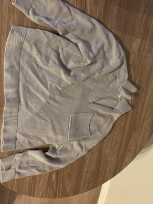 Ljusgrå stickad tröja med halv dragkedja från Arket - Ljusgrå stickad tröja med halv dragkedja vid halsen och en bröstficka. Tröjan har ribbade muddar vid ärmslut och nederkant, samt en avslappnad passform. Perfekt för lager på lager och enkel att matcha.