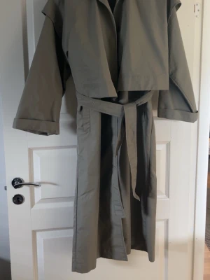Beige trenchcoat med knytskärp XS - Stilren beige trenchcoat i klassisk modell med knytskärp i midjan och bred krage. Jackan har långa ärmar med uppvikta muddar och är tillverkad i ett slätt tyg som ger en snygg siluett. Perfekt för dig som vill ha en tidlös och trendig look.
