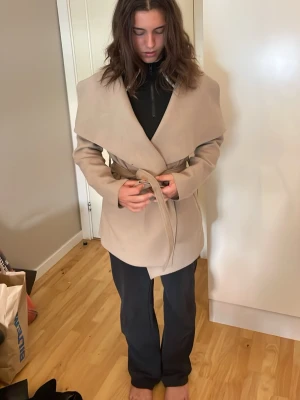 Beige kappa med knytskärp - Stilren beige kappa med bred krage och knytskärp i midjan. Kappan har en avslappnad passform och är tillverkad i ett mjukt tyg som ger en elegant look. Perfekt för dig som vill ha en trendig och enkel ytterjacka till hösten eller våren.