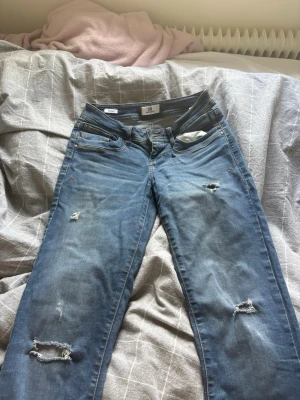 Blå slitna valerie jeans från LTB - Säljer ett par blå jeans från LTB med slitningar och hål på flera ställen för en trendig look. Klassisk femficksmodell med normal midja och raka ben. Jeansen är gjorda i mjukt denimtyg och har snygga detaljer på bakfickorna. Storlek 26/34