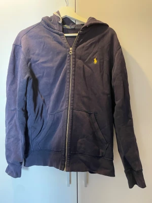 Mörkblå zip hoodie från Polo Ralph Lauren - Mörkblå hoodie med dragkedja från Polo Ralph Lauren. Klassisk gul logga broderad på bröstet, kängurufickor och justerbar huva med snören. Tillverkad i mjukt bomullsmaterial och har ribbade muddar vid ärmar och nederkant. Perfekt för en avslappnad och stilren look.