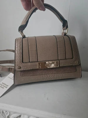 Ny väska - Snygg beige handväska med krokodilmönstrad struktur och guldfärgade metalldetaljer. Väskan har både handtag och axelrem samt ett stilrent lås framtill. Perfekt accessoar för att lyfta din outfit.
