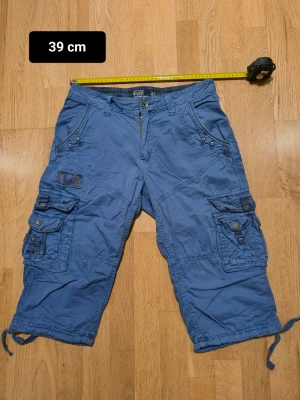 Polo Ralph Lauren Blå cargo shorts med fickor - Säljer ett par blå cargo 3 kvarts shorts med flera stora fickor på sidorna och bak. Byxorna har dragsko vid bensluten och knappgylf. Perfekta för dig som gillar en avslappnad och funktionell stil. Ej använda många gånger 