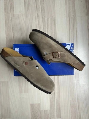 Beige mockatofflor från Birkenstock - Säljer ett par snygga beige tofflor från Birkenstock i mocka med justerbart spänne och korksula. Yttersulan är svart och ger bra grepp. Perfekta för dig som gillar stilren och bekväm design. Klassisk slip-in modell med präglad logga på sidan.