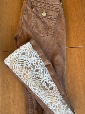 Bruna jeans med spetsdetalj - Unika bruna jeans från True Religion med snygg vit spets längs hela framsidan av benen. Modellen har en bakficka med lock och knapp, och cool tvättad look. Perfekta för dig som vill sticka ut med en kreativ och trendig stil. Sytt bootcut själv