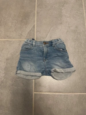 Jeansshorts från Lindex baby - Säljer ett par ljusblå jeansshorts från Lindex för baby, storlek 80. Shortsen har uppvikta benslut, resår i midjan och klassisk femficksdesign. Perfekta för varma dagar och enkla att matcha med olika plagg.