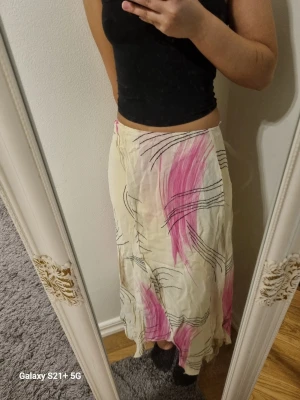 Långkjol med rosa och svart mönster - Säljer en flowy långkjol i ljusbeige med abstrakta rosa och svarta mönster. Kjolen har en luftig passform och är perfekt för dig som gillar unika prints och en bohemisk vibe. Materialet känns lätt och skönt mot huden.
