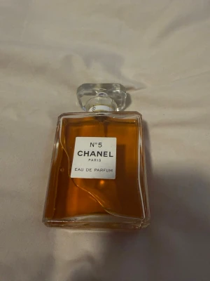 Chanel No 5 Eau de Parfum - Klassisk Chanel No 5 Eau de Parfum i en elegant, rektangulär glasflaska med genomskinlig, multifacetterad kork och guldig detalj. Parfymen har en varm bärnstensfärgad vätska och ikonisk vit etikett med svart text. Perfekt för dig som älskar tidlös och lyxig doft.