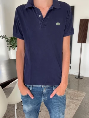 Marinblå pikétröja från Lacoste - Klassisk marinblå pikétröja från Lacoste med korta ärmar och krage. Tröjan har den ikoniska gröna krokodilloggan på bröstet och är gjord i bomull för en skön och luftig känsla. Perfekt till jeans eller shorts för en clean look.