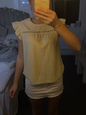 Gul blus med volangärmar - Söt gul blus med broderade detaljer och volang vid axlarna. Blusen har rund halsringning och ett dekorativt hålmönster längs bröstet. Perfekt till sommaren och ger en fräsch look till din outfit. Aldrig använd 