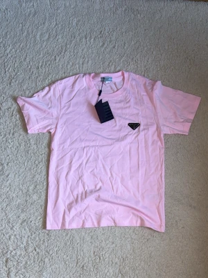 Ljusrosa t-shirt med logga - Snygg ljusrosa t-shirt med korta ärmar och rund hals. På bröstet sitter en liten svart logga som ger en clean och trendig look. Perfekt till jeans eller shorts för en avslappnad stil.