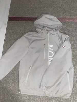 Ljusgrå vindjacka Moncler. - Snygg ljusgrå vindjacka från Moncler med huva och dragkedjor på fickor och bröst. Jackan har Moncler-logga på ärmen och stort vitt tryck framtill. Perfekt för blåsiga dagar och enkel att matcha med olika outfits. Storlek M