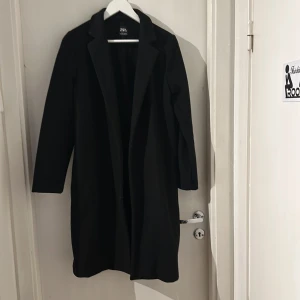 Svart kappa från Zara - Stilren svart kappa från Zara med klassisk krage och lång passform. Jackan har långa ärmar och en rak siluett som ger en clean look. Perfekt för dig som gillar minimalistisk stil och vill ha en tidlös jacka i garderoben. Passar från Stl S-L