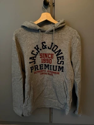 Grå Jack & Jones hoodie med tryck - Säljer en grå hoodie från Jack & Jones med stor tryckt logga och text i marinblått och rött på bröstet. Klassisk känguruficka framtill och justerbar huva med snören. Mjuk bomullsblandning som är skön att ha på sig. Perfekt för en avslappnad stil.