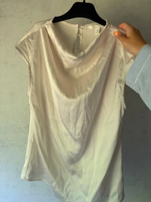 Champagnefärgad satintopp - Snygg topp i champagnefärgad satin med draperad halsringning och ärmlös design. Perfekt för dig som gillar en stilren och trendig look. Toppen har en mjuk glans och faller fint mot kroppen.