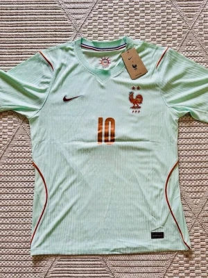 Frankrike fotbollströja Mbappé #10 - Ljusgrön fotbollströja med bronsfärgade detaljer, nummer 10 och Mbappé på ryggen. Tröjan har FFF-logga och Nike-märke på bröstet samt tunna röda linjer längs sidorna. Perfekt för dig som älskar fotboll och vill stötta Frankrike.