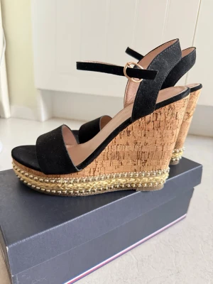 Svarta kilklackade sandaler med pärldetaljer (strl.38) - Snygga sandaler med svart mockaimitation och breda remmar. Kilklack i kork med dekorativa pärlor och flätad detalj runt sulan. Justerbart spänne vid ankeln för perfekt passform. Perfekta för sommarens alla outfits.