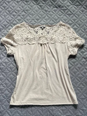 Beige topp med spetsdetaljer - Snygg beige topp med korta ärmar och spetsdetaljer över axlar och bröst. Toppen har rund halsringning och är perfekt för dig som gillar en feminin och stilren look.