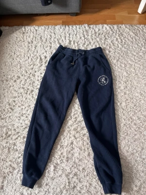 Mörkblå ribbade mjukisbyxor - Mörkblå mjukisbyxor med ribbad struktur och dragsko i midjan. Byxorna har fickor och en vit broderad logga på vänster lår med texten 'RS Deux Ligne New Age Essentials'. Perfekta för chill dagar och streetwear-vibe.