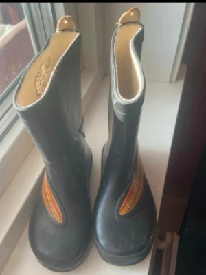 Svarta gummistövlar med färgglada detaljer(barn) - Ett par svarta gummistövlar med insida i beige och dekorativa ränder i orange, rött och gult på ovansidan. Stövlarna har öglor upptill för enkel påtagning och är perfekta för regniga dagar eller festivaler.