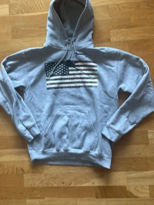 Grå Ralph Lauren hoodie med amerikansk flagga - Säljer en grå Ralph Lauren hoodie med tryck av en amerikansk flagga på bröstet och coolt splattermönster. Tröjan har huva med dragsko och en stor magficka. Perfekt för dig som gillar streetwear och vill ha en avslappnad stil.