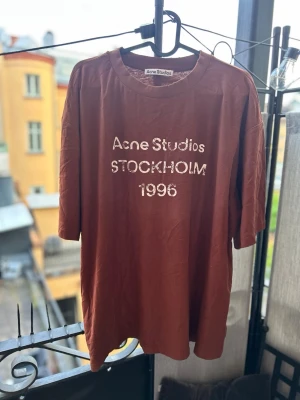 Brun Acne Studios t-shirt - Säljer en brun t-shirt från Acne Studios med trycket 'Acne Studios STOCKHOLM 1996' på bröstet. T-shirten har rund hals och korta ärmar, samt en loose passform som ger en avslappnad vibe. Perfekt för dig som gillar streetwear och enkel stil.