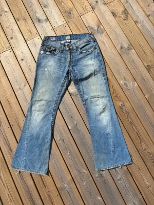 True Religion bootcut jeans - Joey Super T - Säljer dessa feta True Religion jeans i mycket bra skick. Storlek 31. Är en knapp som saknas så satt ditt en annan ( se bild 4) men är inget som syns vid användning. Hör av er vid frågor eller minsta fundering 🙌  Ytterbenslängd: 103 cm  Innerbenslängd: 75 cm  Midja: 38 cm  Benöppning: 25,5 cm