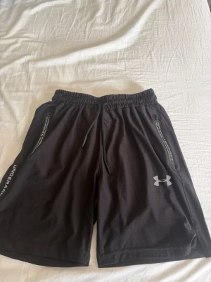 Svarta träningsshorts från Under Armour - Säljer ett par svarta träningsshorts från Under Armour med elastisk midja och snörning. Shortsen har dragkedjeförsedda fickor på sidorna och logga på vänster ben. Tillverkade i ett lätt och ventilerande syntetmaterial, perfekta för träning.