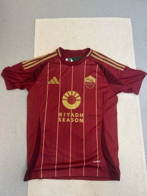 Vinröd AS Roma matchtröja oanvänd med lapp kvar  - Säljer en vinröd AS Roma matchtröja från Adidas med tunna guldiga ränder och detaljer. Tröjan har klubbmärke och sponsorlogga i guld på bröstet samt klassiska Adidas-ränder på axlarna. Tillverkad i ett lätt och ventilerande funktionsmaterial. Oanvänd med lapp kvar 