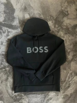 Boss hoodie svart - Snygg svart hoodie från BOSS med stort vitt BOSS-tryck på bröstet. Hoodien har huva med dragsko och en klassisk känguruficka framtill. Tillverkad i mjukt material och har ribbade muddar vid ärmar och nederkant. Perfekt för en avslappnad och trendig look.