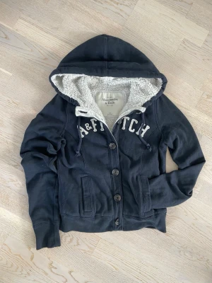 Marinblå hoodie Abercrombie & Fitch - Mysig marinblå hoodie från Abercrombie & Fitch med knappar framtill och vit teddyfoder i huvan. Broderad text 'A&FITCH' på bröstet och två fickor fram. Perfekt för dig som gillar en avslappnad och stilren look. Finns lite fläckar inuti hoodien och en halv knapp saknas ner till.