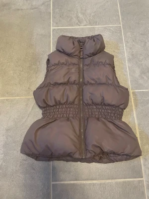 Lila  puffig dunväst med hjärtdetalj - Snygg lila dunväst med hög krage och dragkedja framtill. Västen har puffig design, markerad midja med resår och en söt hjärtdetalj på ryggen. Perfekt för lager på lager och ger en trendig siluett.