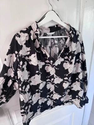 Svart blommig blus från H&M - Snygg svart blus med ljusa blommor och v-ringad hals. Blusen har långa ärmar och ett avslappnat fall. Perfekt för dig som gillar en trendig och bekväm stil. Materialet känns mjukt och lätt mot huden.