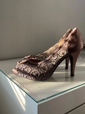 Bruna spetsiga pumps med rosett - Säljer ett par bruna pumps med öppen tå och hög klack. Skorna har ett elegant spetsmönster med blommor på sidorna och en söt rosett framtill. Materialet är en mix av satin och spets som ger en lyxig känsla. Perfekta för dig som vill sticka ut lite extra.