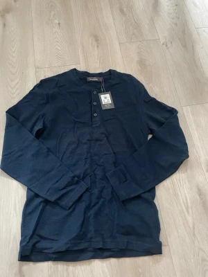 Marinblå långärmad topp Dressmann - Snygg marinblå långärmad topp från Dressmann med knappar framtill och rund hals. Tillverkad i mjuk bomull med diskret ribbad struktur. Enkel och stilren modell som passar till det mesta. Helt orörd