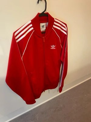Röd Adidas track jacket - Snygg röd track jacket från Adidas med vita klassiska ränder på axlarna och dragkedja framtill. Tröjan har ståkrage, fickor på sidorna och Adidas-logga på bröstet. Perfekt för en sportig och retro look.