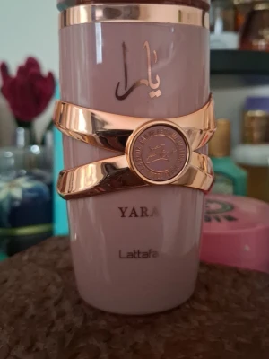 Yara Elixir  - Säljer en elegant parfymflaska från Lattafa, modellen Yara. Flaskan är ljusrosa med guldfärgade detaljer och arabiska bokstäver på framsidan. Designen är lyxig med en unik guldfärgad dekoration runt flaskan.