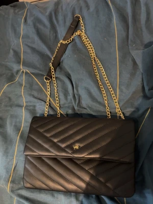 Svart quiltad kuvertväska med kedja - Snygg svart kuvertväska i quiltat mönster med guldfärgad kedjerem och magnetlås. Väskan har en stilren design och ett litet metallmärke på framsidan. Perfekt att bära över axeln med den långa kedjan. Materialet ser ut att vara konstläder.