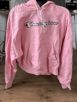 Rosa hoodie från Champion - Säljer en ljusrosa hoodie från Champion med stor logga på bröstet. Hoodien har huva med vita snören och en stor magficka framtill. Tillverkad i mjukt bomullsmaterial och har ribbade muddar vid ärmar och nederkant. Perfekt för en avslappnad och sportig stil.