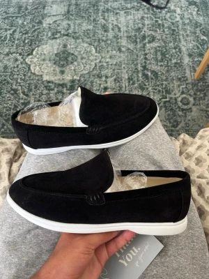 Svarta loafers i mocka - Aldrig använda, Säljer ett par stilrena svarta loafers i mocka med vit sula. Skorna har en klassisk, minimalistisk design utan snörning och passar perfekt till både jeans och chinos. Materialet är mjukt och ger en lyxig känsla. Perfekta för dig som gillar en clean och modern look.