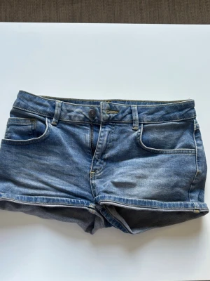 Blå denimshorts med fem fickor - Klassiska blå jeansshorts i denim med fem fickor och uppvikta benslut. Shortsen har normal passform, bälteshällor och knappgylf. Perfekta för varma dagar och enkla att matcha med olika stilar.