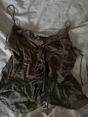 Brun linnetopp med snörning från Zara - Supersöt brun linnetopp från Zara med tunna axelband och snygg snörning framtill. Toppen har volangdetaljer och ett skirt, lätt material som ger en bohemisk vibe. Perfekt för sommardagar och festivaler.