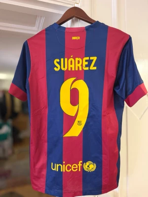 Barcelona matchtröja Suárez #9 - Säljer en officiell FC Barcelona matchtröja från Nike med Suárez och nummer 9 på ryggen. Tröjan har blå och vinröda vertikala ränder, Champions League-märke på ärmen och Qatar Airways som sponsor framtill. Tillverkad i lätt och ventilerande material.