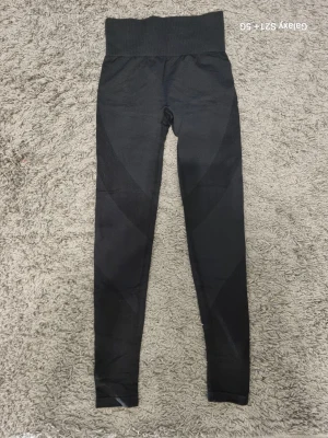 Svarta leggings med ribbade detaljer - Säljer ett par svarta leggings med hög midja och bred linning. De har snygga ribbade partier längs benen som ger en sportig look. Materialet är stretchigt och följsamt, perfekt för träning eller chill. Passformen är tight och benen är långa.