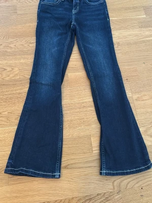 Mörkblå bootcut jeans med vita sömmar och knappfickor i bak - Snygga mörkblå bootcut jeans med klassisk femficksmodell och vita kontrastsömmar. Jeansen har utsvängda ben och knappfickor i bak, perfekta för en trendig och avslappnad stil. Materialet är denim i bomull med lite stretch för extra komfort.